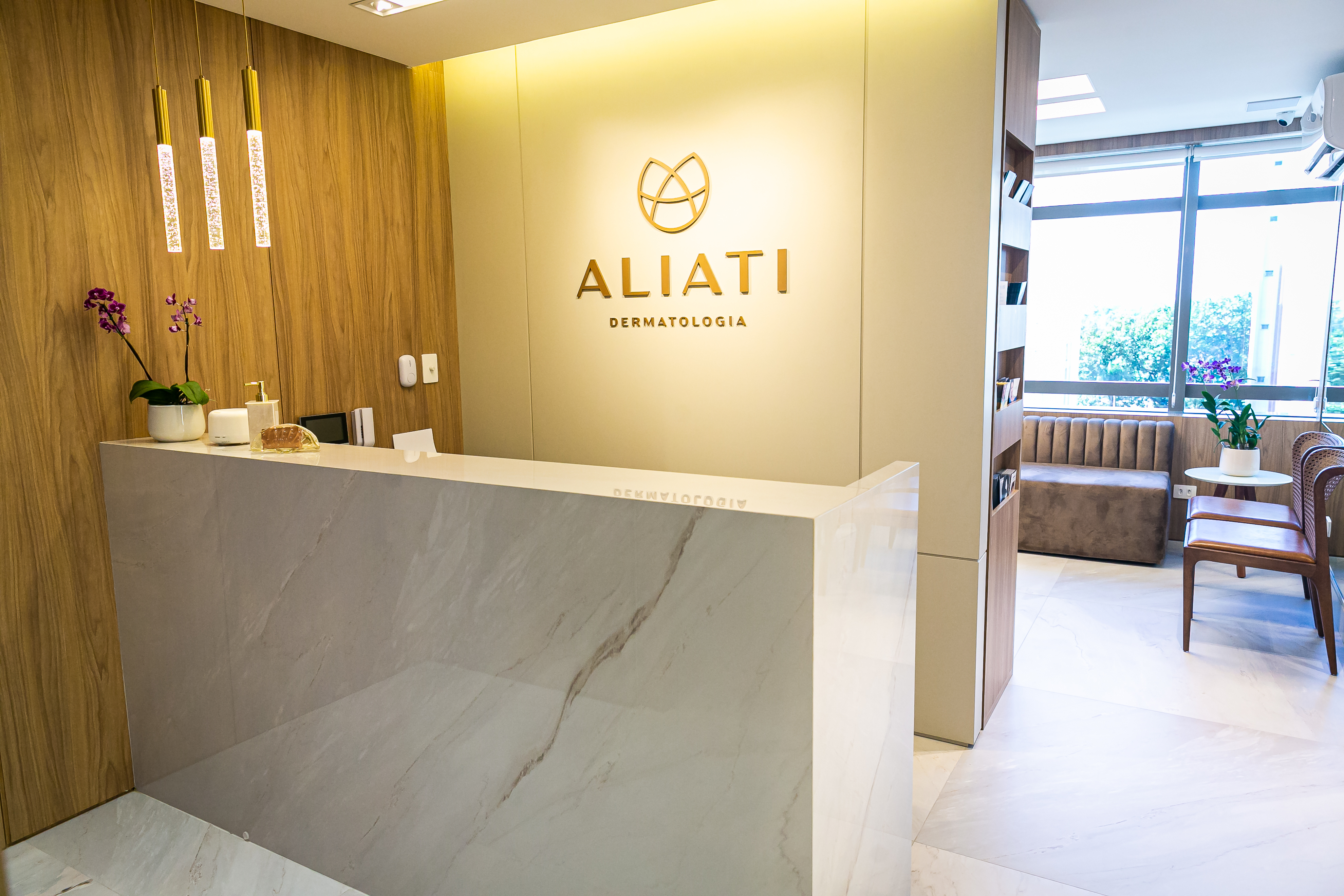 Recepção da clínica à luz quente: parede creme com o logotipo dourado, painel de madeira e balcão com veios de mármore